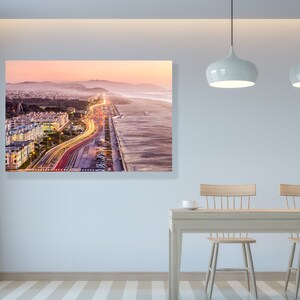 May include: An aerial view of a city at sunset with a highway leading to the beach. The highway is lined with streetlights and the beach is sandy with waves crashing on the shore.