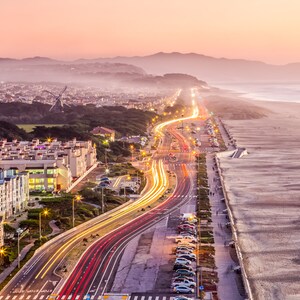 May include: An aerial view of a coastal town at sunset, with a highway lined with streetlights and cars driving along the road. The highway leads to a sandy beach with a seawall and the ocean in the distance.