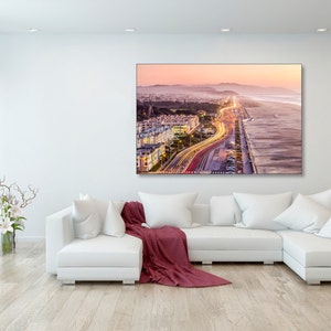 May include: An aerial view of a city at sunset with a highway leading to the beach. The highway is lit up with car headlights, creating a stream of light. The beach is sandy and the ocean is in the background.