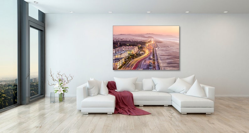 May include: An aerial view of a city at sunset with a highway leading to the beach. The highway is lit up with car headlights, creating a stream of light. The beach is sandy and the ocean is in the background.