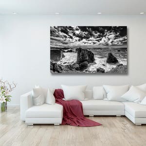 May include: Black and white photograph of a rocky coastline with crashing waves and a dramatic cloudy sky.