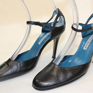 May include: A pair of black leather heeled shoes with a blue lining and a cut-out design. The shoes have a buckle closure and the brand name "Charles David" is printed on the insole.
