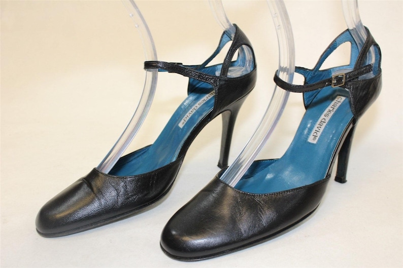 May include: A pair of black leather heeled shoes with a blue lining and a cut-out design. The shoes have a buckle closure and the brand name "Charles David" is printed on the insole.