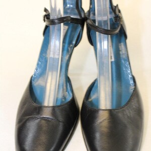 May include: A pair of black leather ankle strap pumps with a low heel. The shoes have a blue lining and are on a white background.