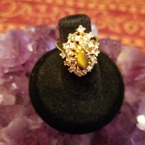 May include: A gold ring with a large, brown tiger's eye stone surrounded by clear rhinestones. The ring is on a black ring display stand.