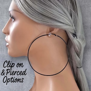 Op de afbeelding: Een vrouw met grijs haar draagt een grote, zwarte oorring. De oorring is bevestigd met een clip. De tekst "Clip on & Pierced Options" is zichtbaar. Het logo van "Hoopaholics" staat rechtsonder.