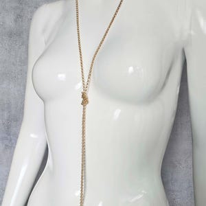 May include: A gold-coloured necklace with a long chain and a small decorative element. The necklace is displayed on a white mannequin. The chain hangs down the front of the mannequin.