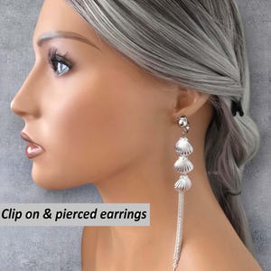 Op de afbeelding: Zilveren oorhangers met een schelpmotief, bestaande uit drie gestapelde schelpen en een franje van bungelende kettingen. De tekst "Clip on & pierced earrings" is zichtbaar.