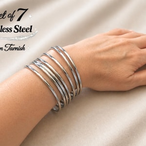 Peut inclure: Ensemble de 7 bracelets joncs en acier inoxydable, non ternissables. Les bracelets argentés sont empilés sur un poignet. Le texte "Set of 7", "Stainless Steel" et "Non Tarnish" est visible.