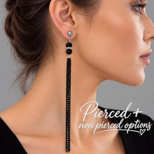 Peut inclure: Une longue boucle d'oreille à pampille noire composée d'un clou argenté, de deux perles noires facettées avec un cristal central et d'une fine frange de chaîne noire texturée. L'image contient le texte Pierced + non pierced options.