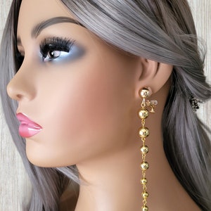 May include: Gold-colored dangle earrings featuring a series of graduated spheres. The earrings are attached to a small gold stud and hang down in a linear fashion. The earrings are shown on a mannequin with gray hair and pink lipstick.
