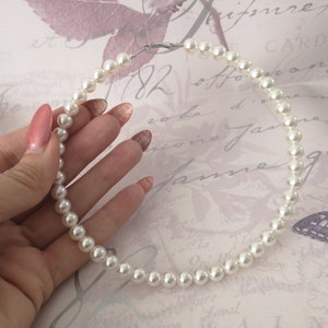 Pu&ograve; includere: Una collana di perle bianche con chiusura argentata. La collana &egrave; tenuta in mano con unghie smaltate. Le perle sono rotonde e ben distanziate, creando un look classico ed elegante. Lo sfondo &egrave; di colore rosa chiaro.