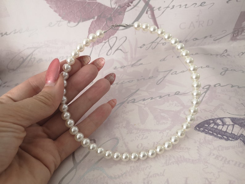 Pu&ograve; includere: Una collana di perle bianche con chiusura argentata. La collana &egrave; tenuta in mano con unghie smaltate. Le perle sono rotonde e ben distanziate, creando un look classico ed elegante. Lo sfondo &egrave; di colore rosa chiaro.