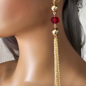 May include: Gold dangle earrings with a red crystal and gold beads. The earrings have a clip-on closure and a long chain tassel.