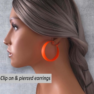 Op de afbeelding: Fel oranje oorringen. De oorbellen zijn rond en hebben een effen kleur. De tekst "Clip on & pierced earrings" staat in een witte doos linksonder.