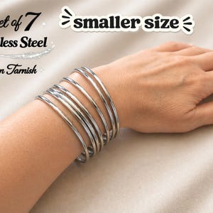 May include: Set of 7 stainless steel bangles in a smaller size. The silver-colored bracelets are non-tarnish and are stacked on a wrist. The image includes text that reads "Set of 7", "Stainless Steel", "Non Tarnish", and "smaller size".