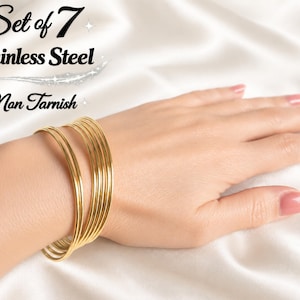 Op de afbeelding: Set van 7 goudkleurige roestvrijstalen armbanden om een pols. De armbanden zijn dun en rond, op elkaar gestapeld. De tekst "Set of 7 Stainless Steel Non Tarnish" staat in de linkerbovenhoek.