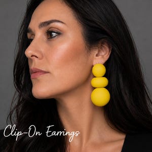 May include: Bright yellow clip-on earrings featuring a vertical arrangement of three smooth, spherical and disc-shaped beads of varying sizes. The text "Clip-On Earrings" is visible in the lower left corner of the image.