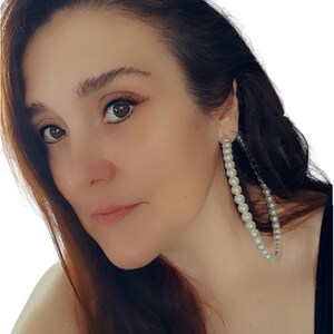 Pu&ograve; includere: Primo piano di una persona che indossa grandi orecchini a cerchio. Gli orecchini sono adornati con molteplici piccole perle bianche rotonde. La persona ha i capelli castani e guarda verso sinistra. Lo sfondo &egrave; bianco.