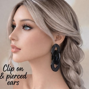 May include: A woman with blonde hair wearing large, black, chain-link earrings. The earrings are oval-shaped and dangle from the ear. The image includes the text "Clip on & pierced ears".