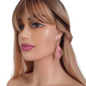 May include: A pair of pink and gold geometric earrings. The earrings are made of thin wire and have a diamond shape. The earrings are hanging from the model's ear.