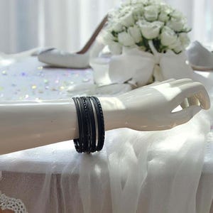 May include: A mannequin arm displays a set of black bangles. The bangles have varying textures, including smooth and patterned surfaces. The background includes a white floral arrangement and a white shoe.