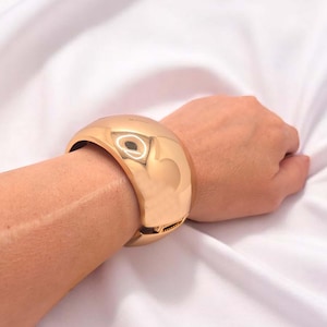 Peut inclure: Un large bracelet manchette de couleur or. Le bracelet est lisse et brillant, reflétant la lumière. Le bracelet est porté au poignet d'une personne. Le bracelet est une pièce audacieuse.