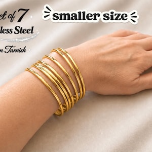 May include: A set of seven gold-coloured stainless steel bangles on a wrist. The bangles are non-tarnish and a smaller size. The bracelets are smooth and shiny, reflecting light. The image includes text that says "Set of 7", "Stainless Steel", and "Non Tarnish".