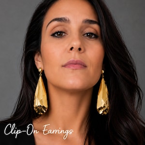 May include: A pair of gold-toned clip-on earrings featuring a faceted, teardrop-shaped design with a polished, reflective surface. The earrings hang from a small, round stud attachment. The text "Clip-On Earrings" is visible in the lower left corner.