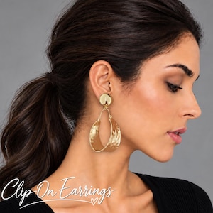 May include: Gold-toned clip-on earrings featuring a smooth, circular top stud connected to a large, textured, open-hoop drop component. The metal surface has a hammered finish that catches the light, creating a reflective and dimensional appearance.