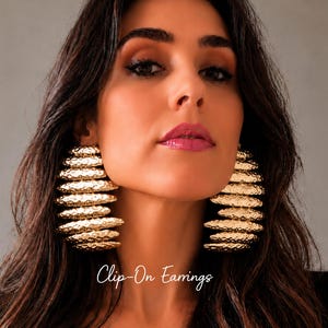 Puede incluir: Un par de pendientes de clip grandes en tono dorado con un diseño apilado de capas horizontales y un acabado texturizado martillado. Los pendientes tienen una apariencia escalonada distintiva que crea un look metálico audaz. El texto Clip-On Earrings es visible.