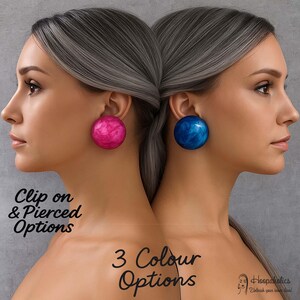 Puede incluir: Dos mujeres con pendientes grandes y redondos. Una lleva un pendiente rosa veteado, la otra uno azul veteado. El texto de la imagen dice "Clip on & Pierced Options" y "3 Colour Options".