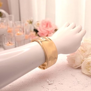 Peut inclure: Un bracelet manchette doré avec un motif texturé est présenté sur un bras de mannequin blanc. Le bracelet a une large bande et une finition lisse et brillante. L'arrière-plan comprend des fleurs et des bougies.