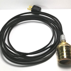 May include: A black pendant light cord with a gold-colored socket and a clear plastic lamp holder. The cord has a black plug and is coiled. The socket is marked with text.