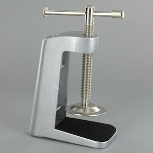 May include: Silver metal clamp with a black rubber base and a threaded screw with a handle. The clamp is designed to attach to a desk or table.