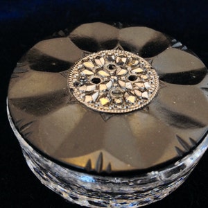 May include: A round, black, cut-glass container with a decorative silver-toned metal embellishment on the lid. The lid has a floral design and the container's base is clear, with a faceted pattern. The container is likely for jewelry or small items.