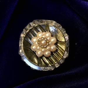 Puede incluir: Broche vintage con diseño floral de perlas de imitación. La flor está centrada en una base oscura, posiblemente de metal, rodeada por un marco circular claro y facetado. El broche probablemente está hecho de metal y vidrio.