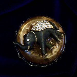 May include: A vintage elephant brooch. The dark gray elephant has gold accents and a jeweled blanket. The brooch is set on a gold-toned, circular base. The background is a dark blue.