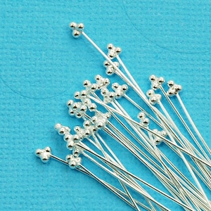 May include: A pile of silver-toned head pins with small round heads. The pins are arranged in a random pattern on a blue background.
