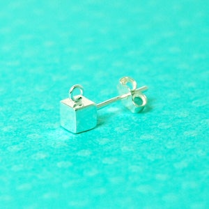Puede incluir: Un pendiente de plata con un pequeño charm de forma cuadrada. El charm tiene un acabado liso y pulido y está unido a un poste con un cierre de mariposa.