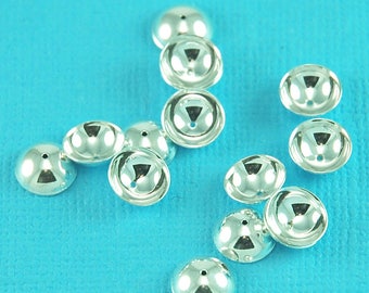 May include: A group of silver-colored metal bead caps scattered on a blue surface. The caps are round and have a domed shape.