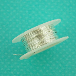 May include: A spool of silver-colored wire on a white plastic spool. The spool is on a turquoise background.