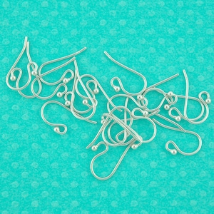 May include: A close-up of a pile of silver earring hooks. The hooks are made of metal and have a small loop at the top. The hooks are arranged in a random pattern on a turquoise background.