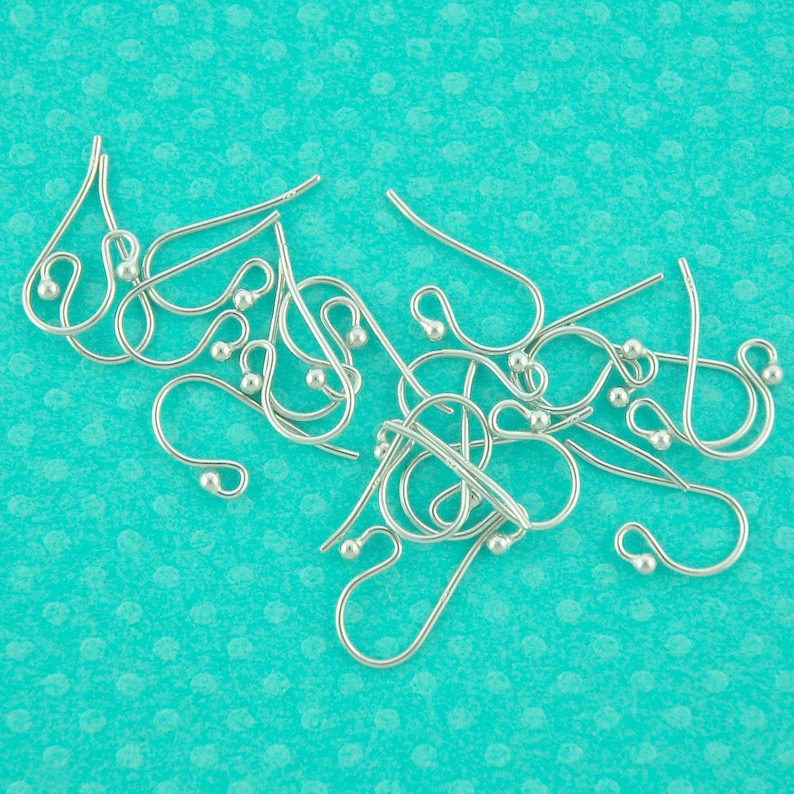 May include: A close-up of a pile of silver earring hooks. The hooks are made of metal and have a small loop at the top. The hooks are arranged in a random pattern on a turquoise background.