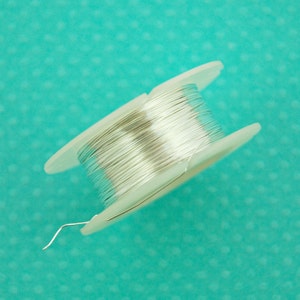 Puede incluir: Una bobina de alambre plateado en una bobina de plástico blanco. El alambre está enrollado apretado y está listo para ser utilizado para manualidades o la fabricación de joyas.
