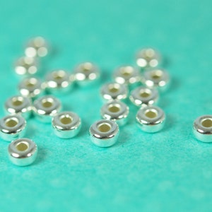 May include: A close-up of silver metal beads scattered on a turquoise surface. The beads are round and have a smooth, shiny finish.