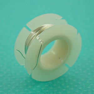 May include: A spool of silver wire on a white plastic bobbin. The wire is wound around the bobbin and the end of the wire is sticking out.