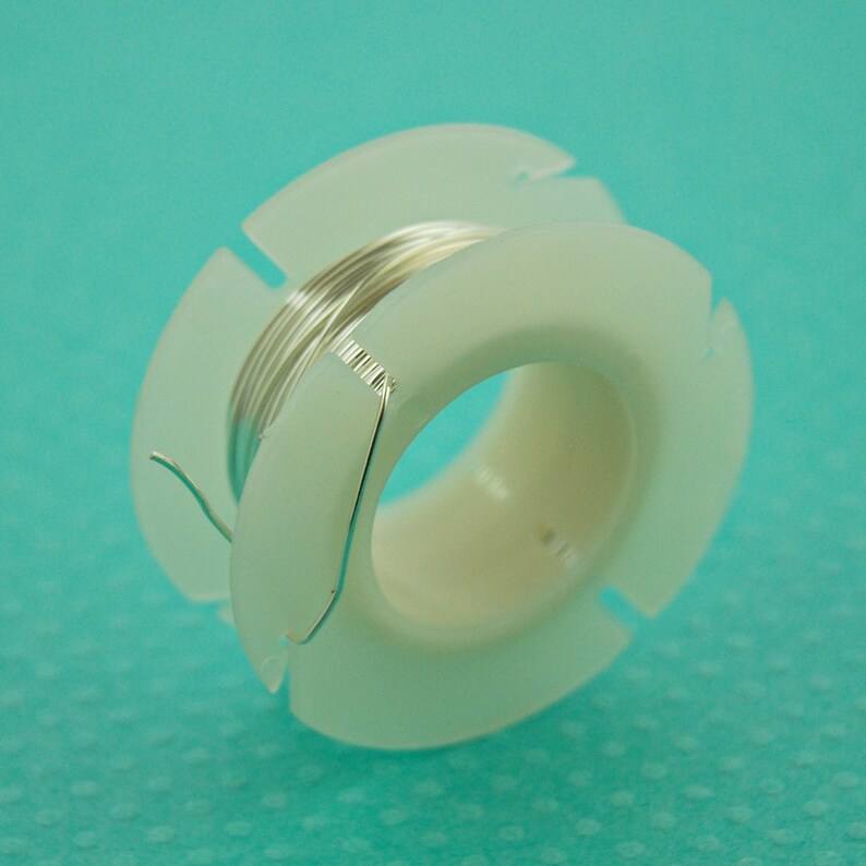 May include: A spool of silver wire on a white plastic bobbin. The wire is wound around the bobbin and the end of the wire is sticking out.