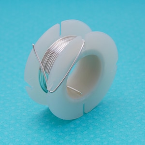 Può includere: Un rotolo di filo d'argento su un distributore di plastica bianca. Il filo è avvolto strettamente attorno al rotolo.