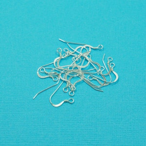 Puede incluir: Un montón de ganchos para pendientes de plata sobre un fondo azul. Los ganchos son de metal y tienen un diseño sencillo.